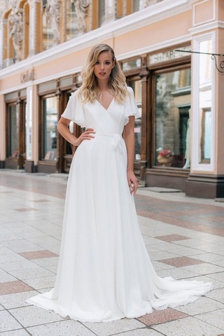 Blondes Model in einem weißen Chiffonbrautkleid mit Flatterärmeln vor einer italienischen Einkaufsstraße von vorne