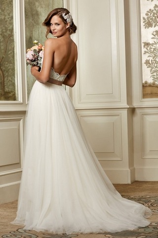 Model mit halblangen Haaren mit einerm weißen Tüllkleid mit braunem Gürtel und einem Blumenstrauß indoor in einem schloßähnlichen Raum von hinten