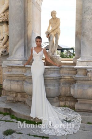 Brünettes Model mit Pferdeschwand in einem körperbetonen weißen Brautkleid aus Georgette mit Spitzenoberteil und Spitze in der Schleppe vor einer antiken mediterranen Kulisse von vorne