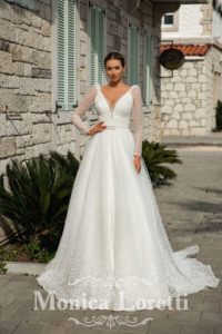 Brünettes Model mit Dutt in einem langen weißen Tüllbrautkleid mit Pünktchen und Puffärmeln in mediterraner Kulisse von vorne