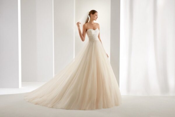 Blondes Model mit Pferdeschwanz mit beigefarbenem Tüllbrautkleid mit Corsage und langem Schleier vor weißbeiger Fotokulisse von vorne