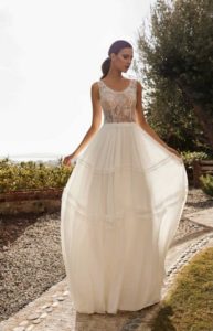 Model mit langen braunen Haaren in einem weißen Bohokleid mit abgesetzten Volants und Spitzenoberteil mit Trägern und Rundhalsausschnitt in einem Park von vorne