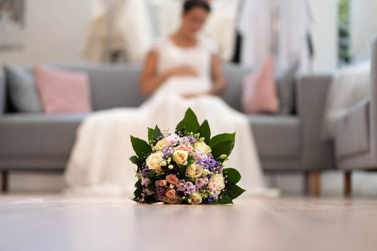 Brautstrauss im Vordergrund, im Hintergrund unscharf schwangere Braut im Brautkleid auf Sofa sitzend