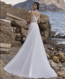 Braunhaariges Model mit Hochsteckfrisur in einem Chiffon-Brautkleid in offwhite mit transparentem Oberteil mit Spitzenapplikationen und Spaghettiträgern an einem Strand von hinten