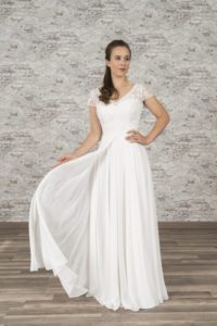 Model mit langen brauenen Haaren in weißem Chiffonbrautkleid mit Bohospitze im Oberteil und kurzen Ärmeln, Rock doppellagig mit Öffnung in der Mitte vor bräunlicher Fotokulisse von vorne