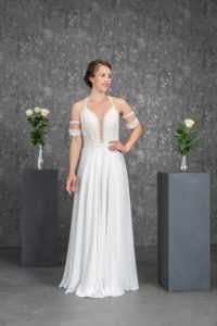 brünette Frau mit Hochsteckfrisur in weißem Chiffonbrautkleid mit raffiniertem, tiefen Ausschnitt in Spitze und kleinen Ärmelchen in Fotokulisse von vorne