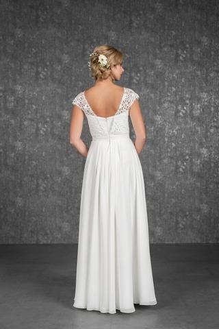 Blondes Model mit einer Steckfrisur mit Blüten in einem Chiffon-Brautkleid in weiß mit Baumwollspitze und leicht überschnittenen Ärmeln vor einer grauen Fotokulisse von hinten