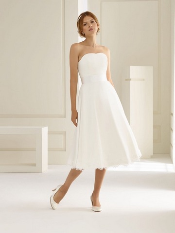 Blondes Modell in einem weißen kurzen Corsagenbrautkleid mit breitem Gürtel vor einer weißen Fotokulisse von vorne