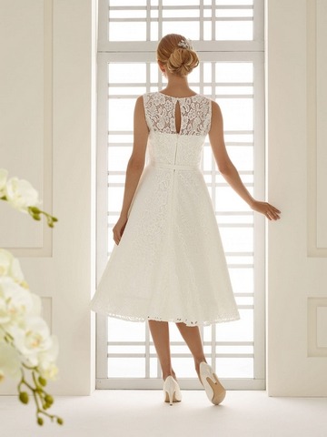Blondes Model mit Dutt in einem weißen schwingenen kurzen Tüllbrautkleid mit Spitzenoberteil ohne Ärmel von hinten in einem weißen Zimmer vor einer Glastür