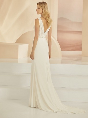 Blondes Model mit offenen Haaren in einem schlichten weißen Brautkleid mit Spitzenoberteil und kleiner Schleppe vor einer weißrosefarbenen Fotokulisse von hinten