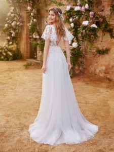 Model mit langenn Haaren in Brautkleid mit Tüllrock, feiner spanischer Spitze mit Flügelämelchen vor Rosenbüschen von hinten