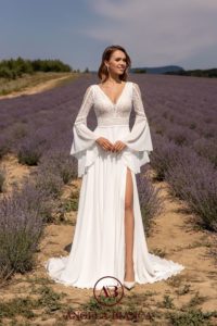 Brünettes Model in einem weißen Boho-Chiffonkleid mit Spitze V-Ausschnitt, weiten langen Trompetenärmeln, und hohem Beinschlitz im Rock im Lavendelfeld stehend, von vorne