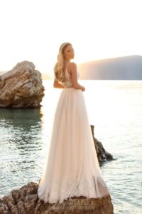 Blondes, langhaariges Model mit weißem Tüllbrautkleid mit Spitzenoberteil und Spaghettiträgern auf einem Fels am Meer stehend von hinten