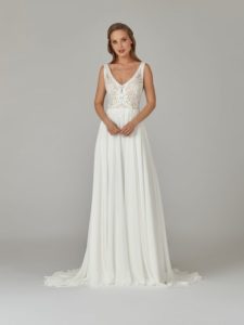 Brünettes Model mit langen offenen Haaren in einem weißen Chiffonbrautkleid mit Boho-Braumwollspitze im Oberteil und Trägern vor einem grauen Fotohintergrund von vorne