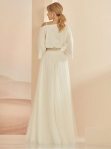 Blondes Model in weißem One-shoulder-Brautpulli, silbernem Grütel und weißem Tüllrock vor rosefarbenem Fotohintergrund von hinten