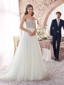 Brünettes Model mit offenen Haaren in einem weißen Corsagenbrautkleid mit Spitzenoberteil und Tüllrock mit Schleppe zusammen mit Bräutigam vor einer gedeckten Tafel von vorne