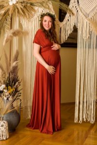 Brünettes, schwangeres Model in einem rostfarbenen langen Wickelkleid mit kurzen Ärmeln vor einer Bohokulisse mit Trockenblumen und Makramee