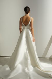 Braunhaariges Model mit Dutt in einem weißen Satinbrautkleid mit breitem Bund, Spitzenoberteil und Trägern und einer Knopfleiste über den ganzen Rock vor einer grauen Fotokulisse von hinten