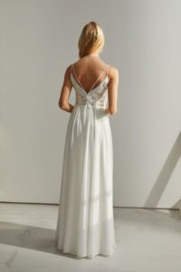 Blondes Model mit einem weißen Bohobrautkleid mit Spitzenoberteil und doppelten Trägern vor einer grauen Fotokulisse von hinten