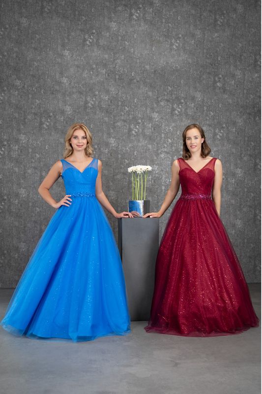 2 Models mit weiten Tüllabendkleidern, eines in royalblau das andere in bordeauxrot. Dazwischen eine graue Stele mit einem Blumentopf mit weißen Blumen vor einem grauen Fotohintergrund