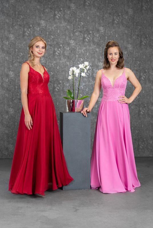 2 Models mit einem roten und einem rosefarbenen Abendkleid in der Mitte eine Steele mit einem Blumentopf und einer weißen Orchidee vor einem grauen Fotohintergrund
